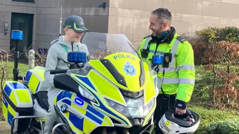 A boy in a grey tracksuit is sitting on a police motorbike while a police officer is stood next to him and smiling at him