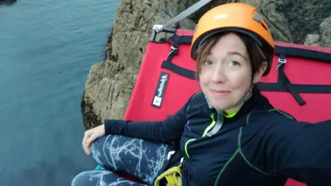 Sally Millington A woman wearing gym clothes and an orange hard helmet camps on the side of a cliff near the sea with a red bed.