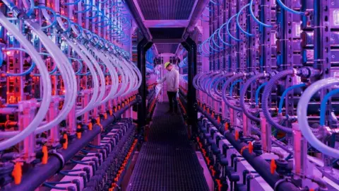 Vaxa A walkway lined with bioreactors emitting pink and purple light. At one end, a worker checks equipment.