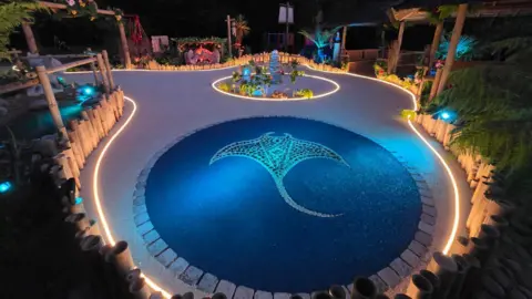 Green Genie Brothers A birds-eye view of a transformed garden at night. There are palm trees and a giant mosaic sting ray in the middle of the garden. It is lit up and in a Hawaiian- style.