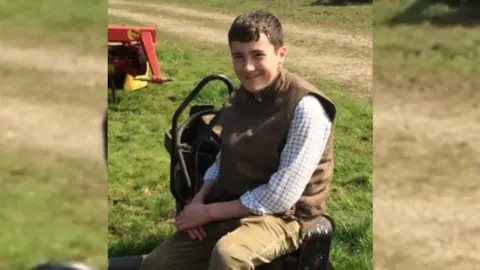 Handout Harry Churchill as a pre-teen with short brown hair with a longer fringe. He is wearing a brown gilet and a white checked long sleeve shirt with light sand coloured corduroy trousers and sitting on a small piece of farming equipment. There is grass in the background and Harry is smiling at the camera. 