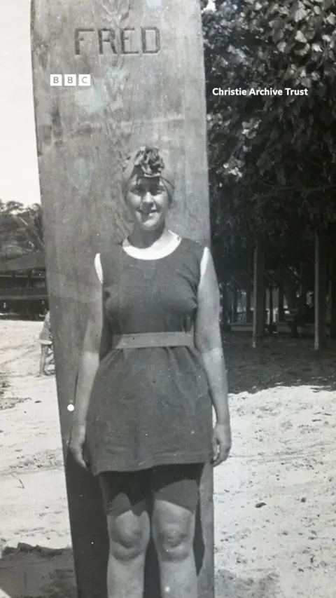 A young Agatha Christie stands on a beach against a large surf board.