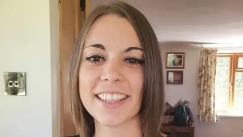 Shannon smiles at the camera. She has a sleek bob haircut in light brown hair. She is standing in the doorway of a room in her house, there are pictures on the wall behind her and a window with curtains.