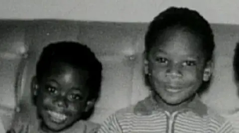 Alder family A black and white photo of Janet and Christopher sitting on a leather sofa when they were children. Janet is on the left wearing a capped-sleeve dress and Christopher is on the right wearing a striped t-shirt