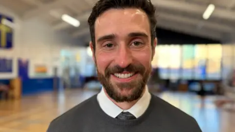 A man with a beard is wearing a dark tie and jumper and a white shirt. He is smiling at the camera.