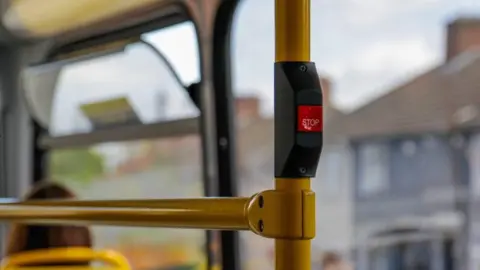 A generic image taken from inside a bus features a stop button. Houses visible through windows in the background are blurred.
