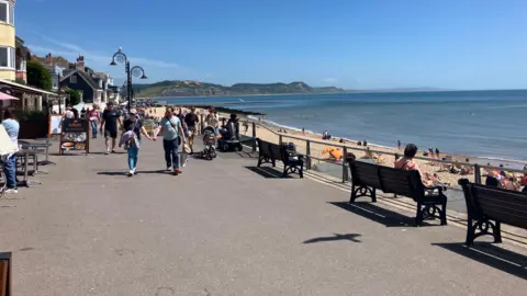 People walking along the promenade in Lyme Regis, with many people sat on the beach or nearby benches enjoying the sun.
