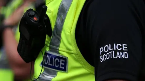 Police Scotland in white bold letters in a black sleeve worn by an officer, with a yellow hi-vis top and walkie-talkie style radio attached.