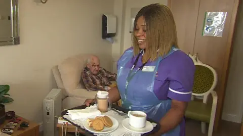 Charity Lawrence, a health care assistant in a care home, wears a purple uniform and holds a tray of tea and biscuits. She has shoulder-length braided hair and is smiling. Behind her is an elderly man in a chair.