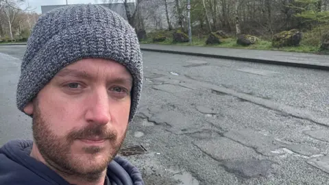 Jamie Lee Collins Man in his late thirties stood outside next to a road. He has a beard and is wearing a light grey beanie hat and a dark grey hoodie.