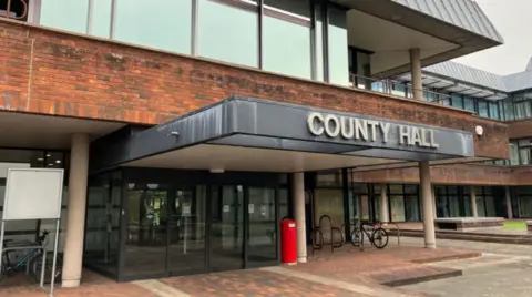 The main entrance to County Hall, the former headquarters of Worcestershire County Council. It is a brick building with a grey canopy at its entrance. The words 'county hall' are attached to the canopy. There are glass automatic doors, with a red bin outside and a bicycle stand nearby.