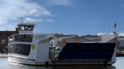 The Floating Bridge 6 crossing the River Medina. It is white, with large blacked out glass panelling and a ramp 