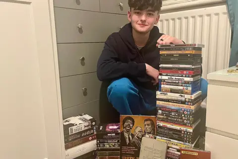 Declan sitting on the floor next to two tall stacks of DVDs and box sets, with additional DVDs arranged in front. The scene includes a grey chest of drawers on the left and a white radiator in the background.