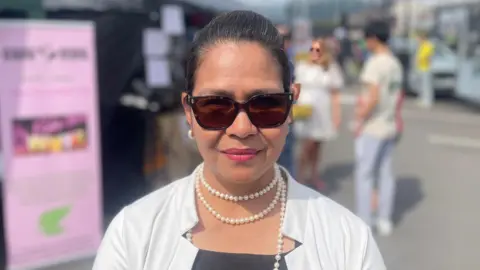 A long brown haired woman wears a white shirt with a black top underneath. She has a pearl necklace around her neck. She wears brown tinted sunglasses and pink lipstick. Behind her, out of focus, are black and green market stalls.