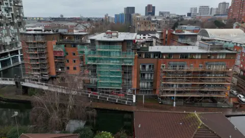 A drone image of the apartment block with has several storeys and lots of windows, however may of these are covered up by metal scaffolding and green netting stretching across it.