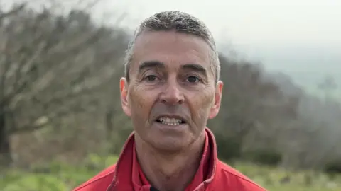 A man with short grey hair and brown eyes is pictured looking directly at the camera wearing a red fleece. He's stood outside with trees and grassland behind him.