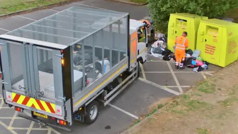 Slough Borough Council A council worker dressed in fluorescent suit standing over fly-tipped rubbish next to two charity bins.