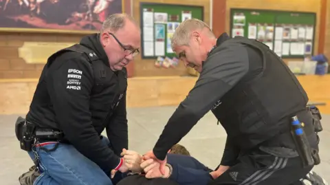 During a police training exercise, officers in casual clothes, with police vests on, are handcuffing another police officer in plain clothes who is lying down on his front with his hands behind his back. They are in a training hall.