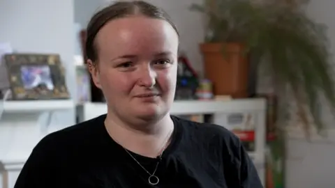 BBC Lily has her brown hair tied up and is looking behind the camera. She's wearing a black T-shirt and a silver necklace. Sat in front of a homely background with a photo frame and plant.