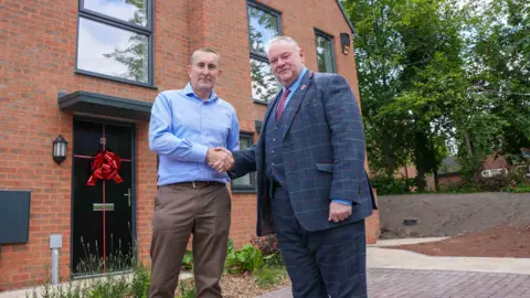 Two men shake hands outside a newly-built house which has a red ribbon tied to a black front door. There are trees and further houses in the background.