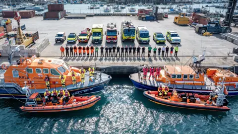 RNLI Various members of emergency services posing on a dock with vehicles and boats 