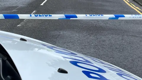 The white bonnet of a police van parked next to a blue and white police cordon stretched across a grey road.