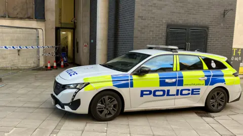 A Suffolk Police car parked in front of a building with blue and white police tape in front of it.