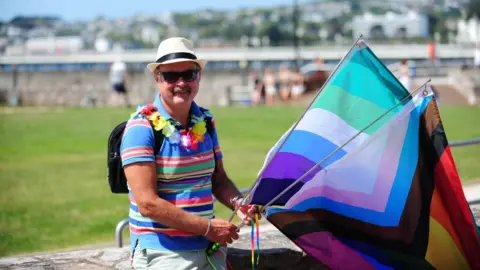 Harry Schofield A man wearing a multicoloured striped top and flower chain around his neck. He is wearing a white hat and black backpack. He is holding two flags, one that combines the trans flag and pride flag and the other a non-binary flag. 