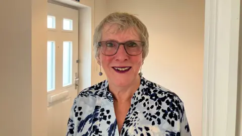 JOANNA TAYLOR/BBC Alison Jones is smiling and looking at the camera. She is wearing glasses, dangly earrings and a pale blue shirt with a darker blue and white spotted pattern. Behind her is a white wall and white door (the inside of the front door).