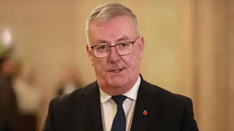 PA Media Mike Nesbitt - a man with grey hair wears square framed glasses as well as a poppy pin, dark suit jacket, a white collared shirt and dark tie is speaking.