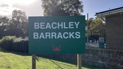 There's large green sign with bold white text that reads “BEACHLEY BARRACKS”. Below the text, there is a red emblem which represents military insignia. The sign is on a neatly mown grassy area with trees with green foliage, a brick wall, which is the entrance to the barracks.