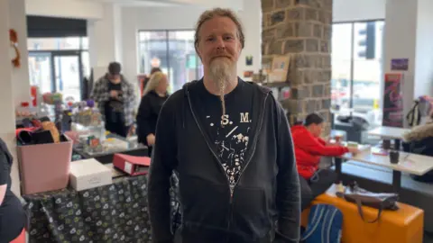 A man with a long grey beard, tied into a plait, is wearing a black hoodie over a black t-shirt as people play board games in the background.