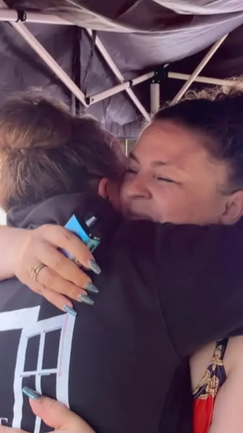 Two people hug closely under a tent frame, one holding a small blue device while wearing a hoodie with a white graphic.