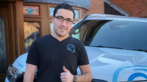 Supplied A man with a black branded T-shirt and glasses giving us a thumbs up with a branded car in the background. There is a house behind the car.