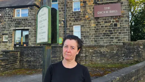 Aisha Iqbal/BBC A woman, dressed in black, stands in front of a pub building, built from heritage stones. It has maroon signs with gold writing identifying it as 'The Dickie Bird'. 