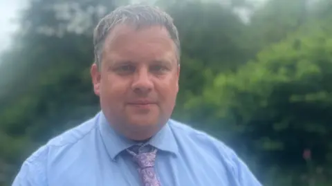 Councillor James Mustoe smiles at at the camera as she stands outside surrounded by trees and hedges. he is wearing a shirt and tie.