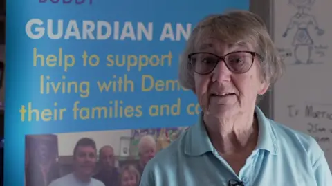 Anne with grey hair wearing a blue shirt with a Good Deeds Trust logo on it.