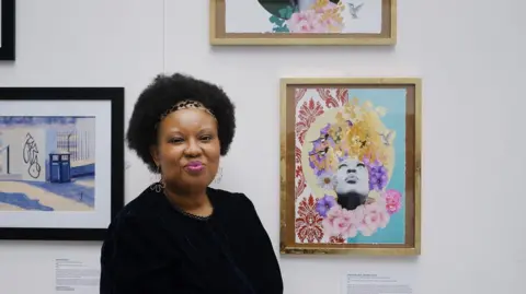 Irina Mackie Sandra Palmer, known as The Brummy Artist, stands next to paintings displayed on a gallery wall.