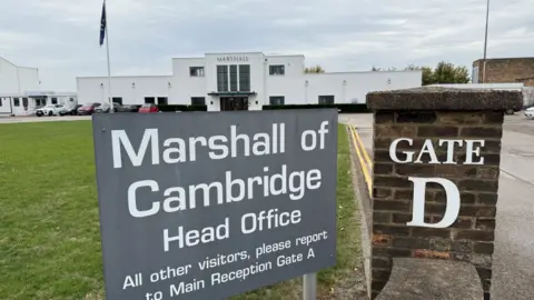The entrance to Marshalls head office. A white building with three large windows at the front. There is a small car park to the front of the building and a sign that reads "Marshall of Cambridge Head Office". 