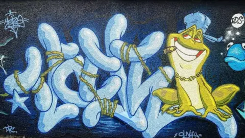 James Thompson/Wall Games A street art mural featuring a frog wearing a hat on sideways and a gold chain round its neck. It is smiling and has a cigar in the side of its mouth. It is sitting on some lettering with some vines weaving around the lettering.