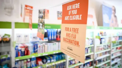 A series of orange and white signs hang on a thread across a pharmacy with the one in the foreground split into a dark orange in the top half and light orange in the bottom half. The text on the top half reads "Ask if you are eligible". The bottom half reads "For a free NHS Flu vaccination" followed by some smaller text below it. On the background are shelves stacked with boxes and bottles of varying colours.