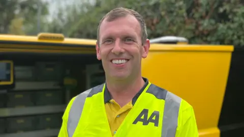 Lee stands smiling next to a yellow van. The door is open and tools are visible inside. He is wearing a high visibility vest that features the "AA" logo.