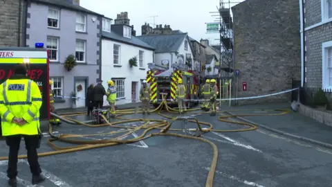 View down a street of firefighters and two appliances, with a tangle of fire hoses on the ground. A police officer stands on the left of the scene,