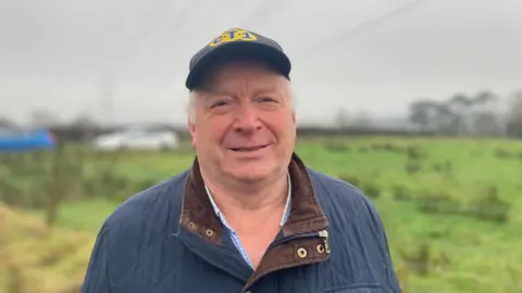 Ian Montgomery. He Has short white hair, wearing a cap with 'C.T.1' on it and a navy jacket. Fields are in the background.