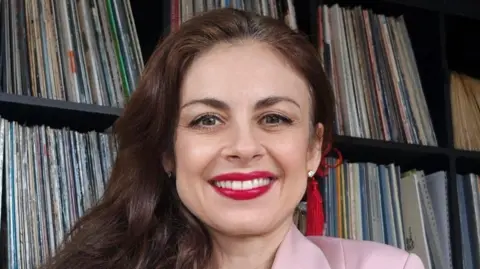 Handout A woman with long, brown hair and red lipstick. SHe is wearing a pink blazer and sitting in front of a shelf of vinyl records.