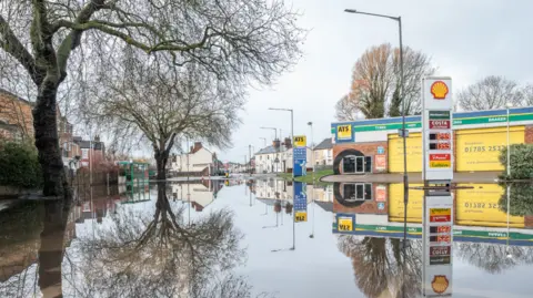 Ian Knight / Z70 Photography A flooded area, with the images of two trees on the left showing in the water. A Shell petrol board and tyre retailer ATS premises are on the right. 