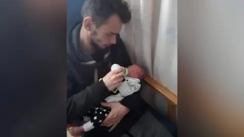North Wales Police Will is seen feeding a bottle to newborn baby Ethan. He is sat down in a chair with a wooden frame with a white curtain on the right hand side. 