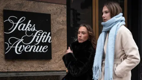 Two women with light brown hair and wrapped in winter jackets walk past a store. A sign on the building reads Saks Fifth Avenue. 