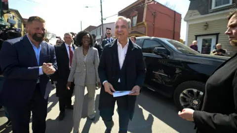 Reuters Mark Carney, in a dark suit and no tie,  smiles as he walks outside with Terrebonne candidate Tatiana Auguste, in a grey pinstripe suit, on a sunny day. 