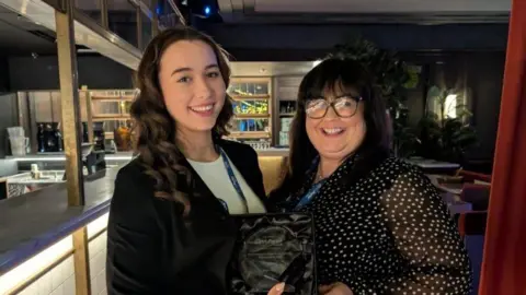 Two women stand in an empty restaurant holding an award. One is younger with long curly brown hair. The older woman has black hair and a fringe and is wearing black-rimmed glasses.